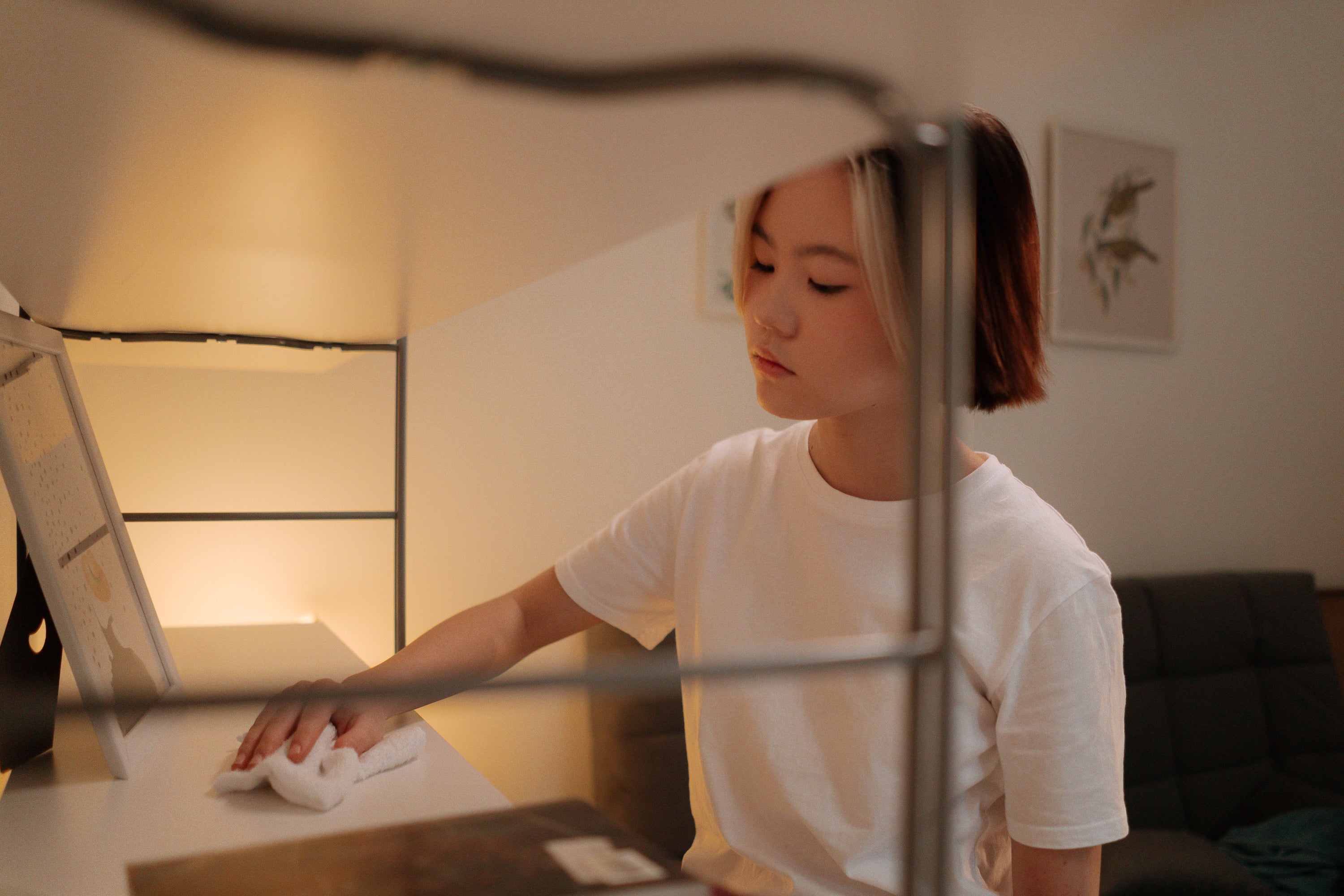 Asian woman in white shirt cleaning shelf indoors with a cloth.